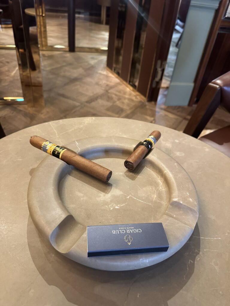 Two cigars rest on a circular marble table in a lounge, next to a navy blue Cigar Club card and ashtray.